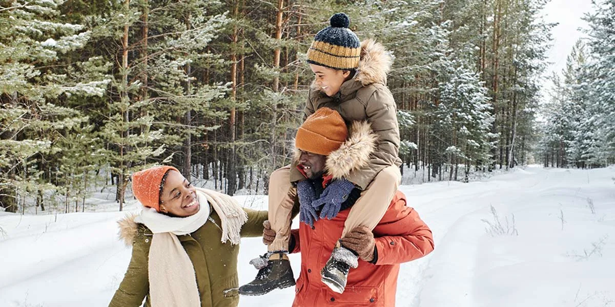 Four people enjoying winter in a snowy forest, with one person giving another a piggyback ride, all dressed warmly in winter clothing.