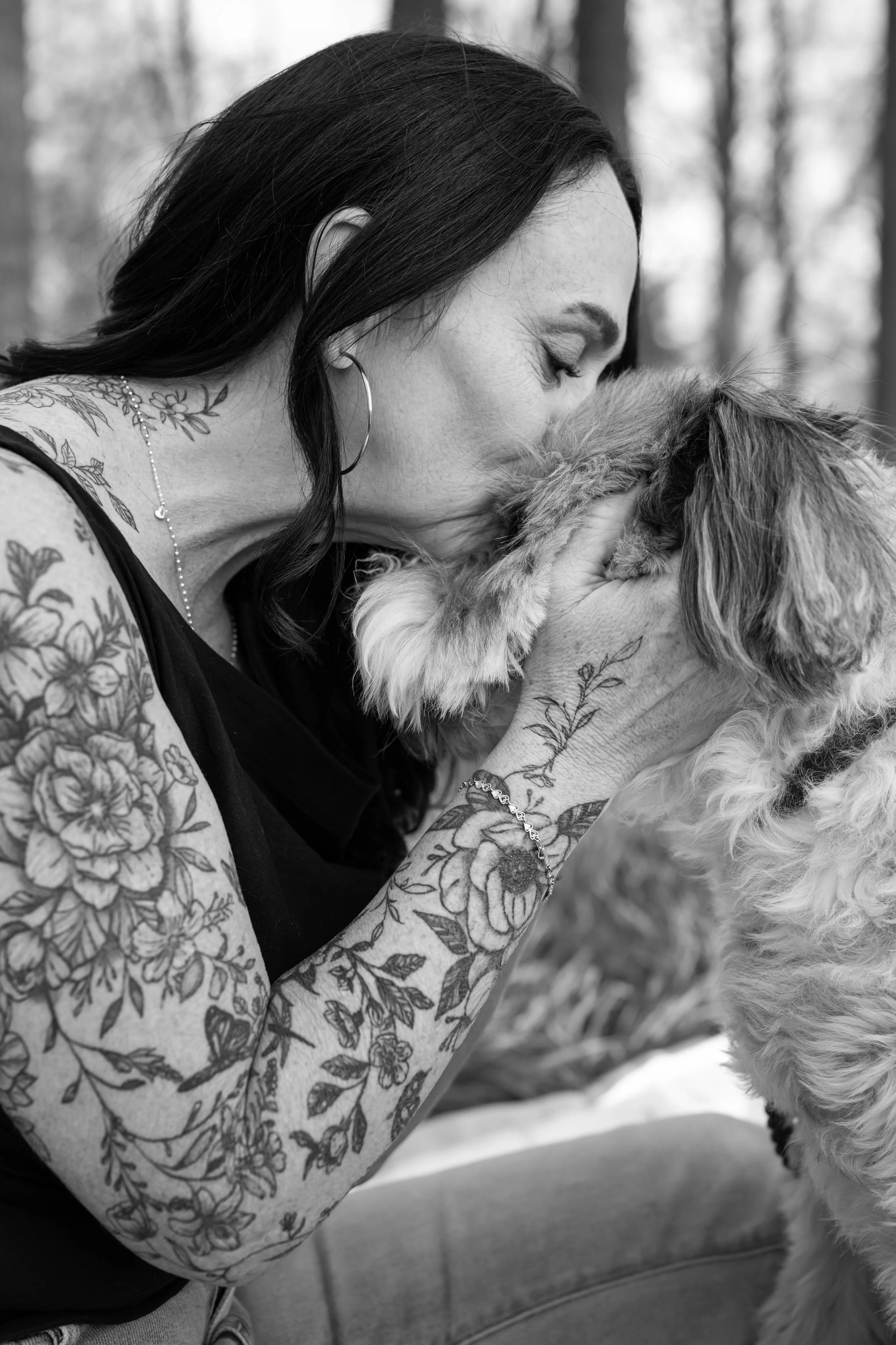 A woman with dark hair and tattoos on her arms and neck is holding and gently kissing a fluffy dog in an outdoor setting with trees in the background.