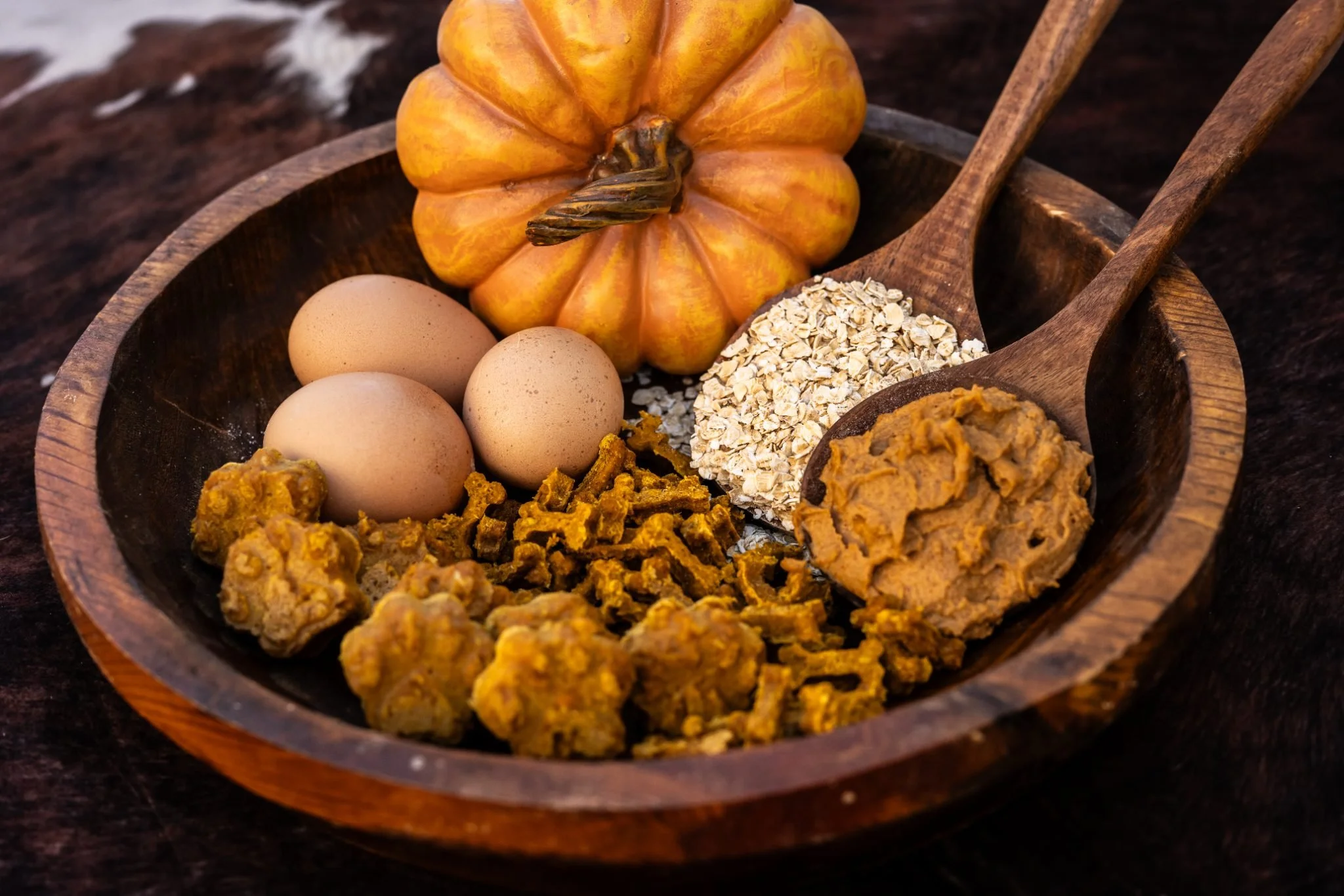 A wooden bowl containing three brown eggs, a small pumpkin, oats, and turmeric cookies, with wooden spoons beside it on a dark surface.