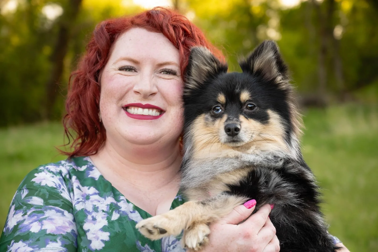 A woman with curly red hair and a bright smile holding a small black and tan dog outdoors with trees in the background.