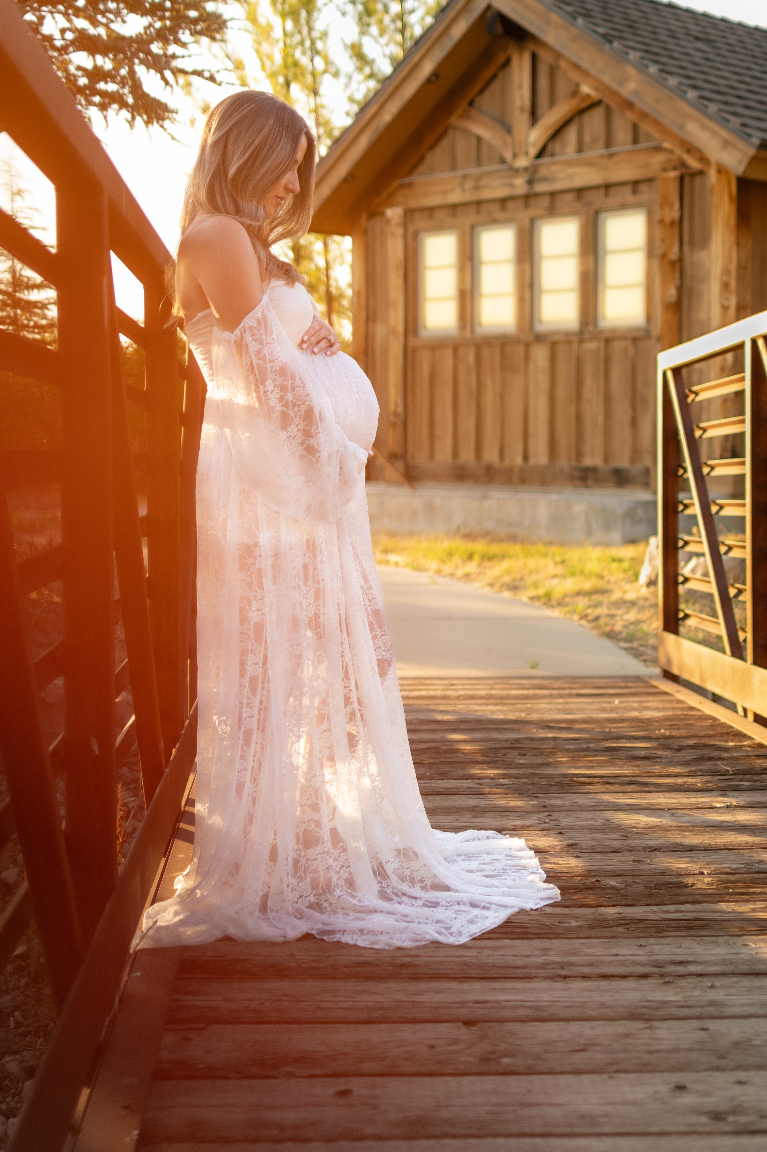 Women holding their pregnancy belly in a flowy lace dress, leaning against the side of a bridge with a wood cabin in the background