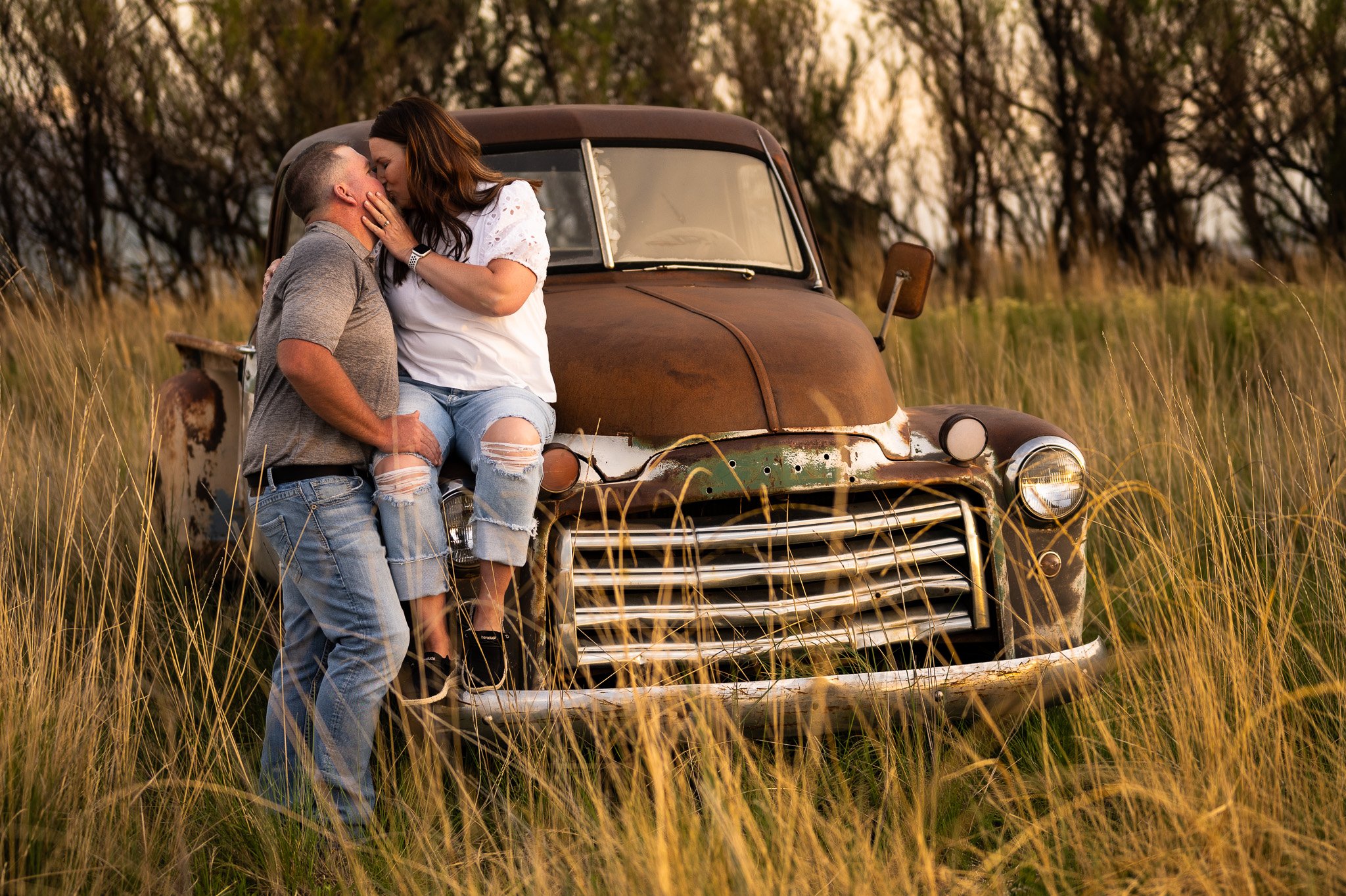 A couple kissing while the female is proped up on the hood of an older looking truck in a field