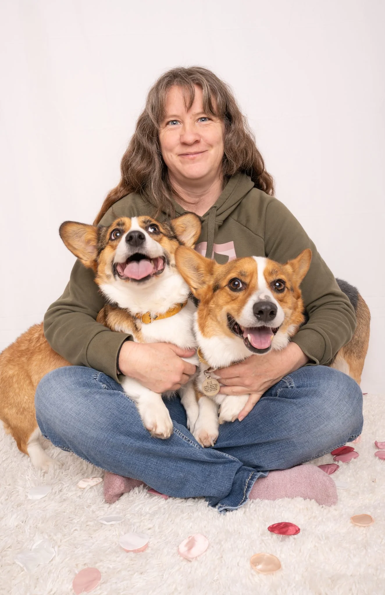 A woman sitting cross-legged on a white fluffy rug, holding two happy corgi dogs, with rose petals scattered on the rug.