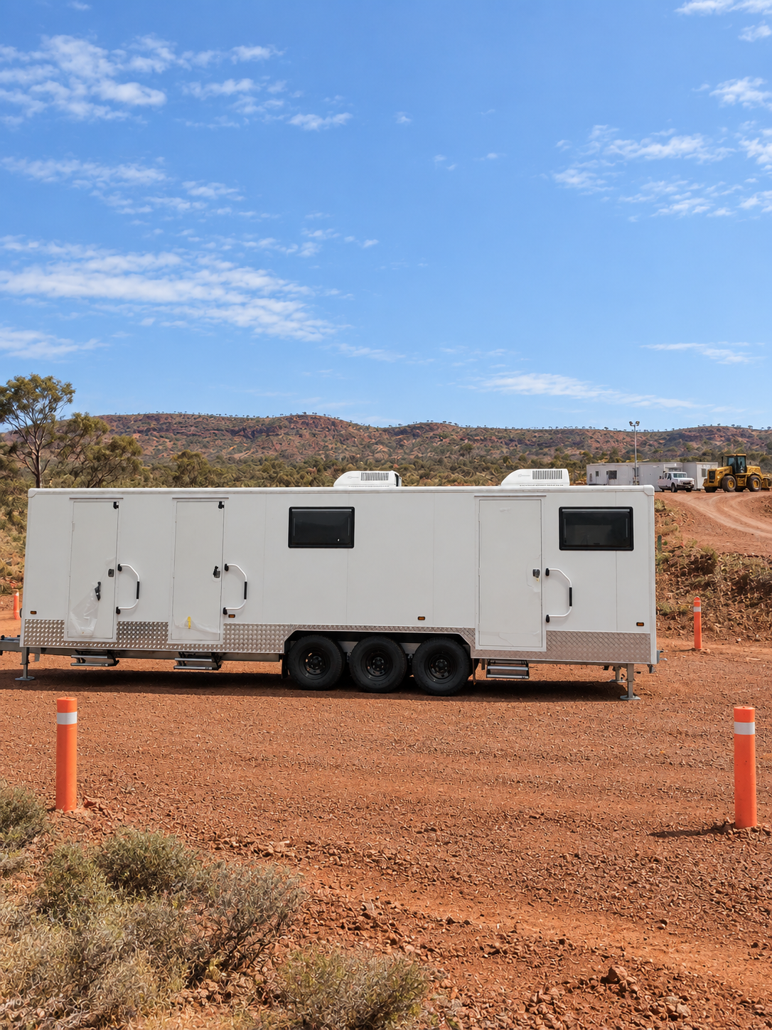 White mobile trailer with three axles parked on reddish dirt ground in a semi-arid landscape, with hills and other trailers in the background under a blue sky with scattered clouds.