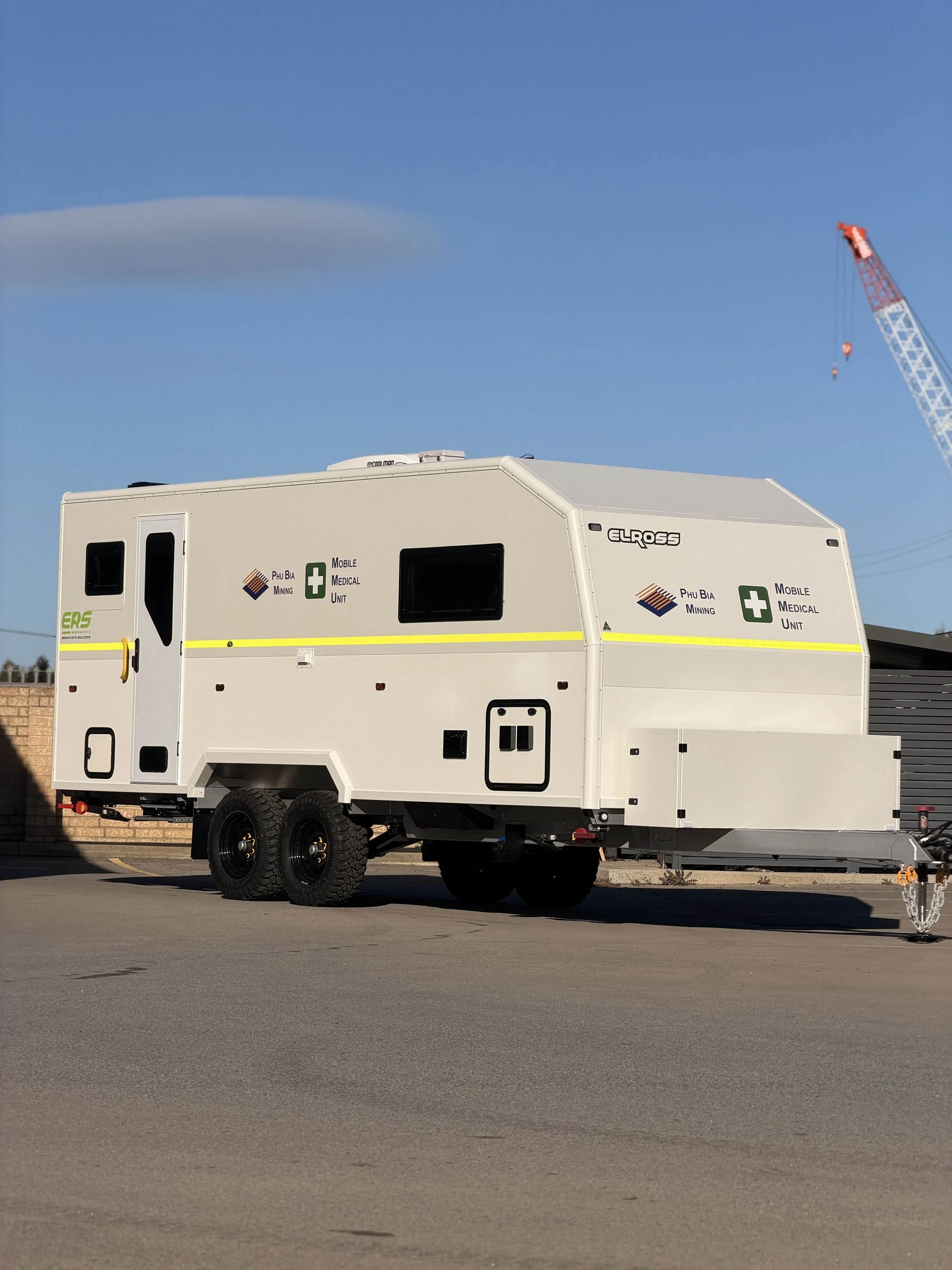 White mobile medical unit trailer with logos and text, parked on a paved lot, with a crane and blue sky in the background.