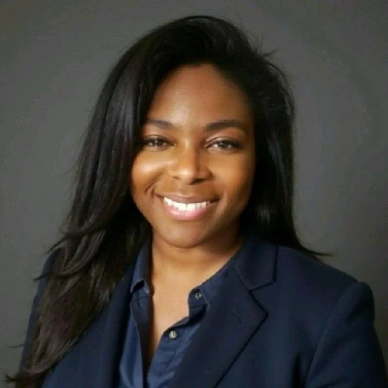 Smiling person with long dark hair and wearing a blazer, in an indoor setting with exposed brick wall.