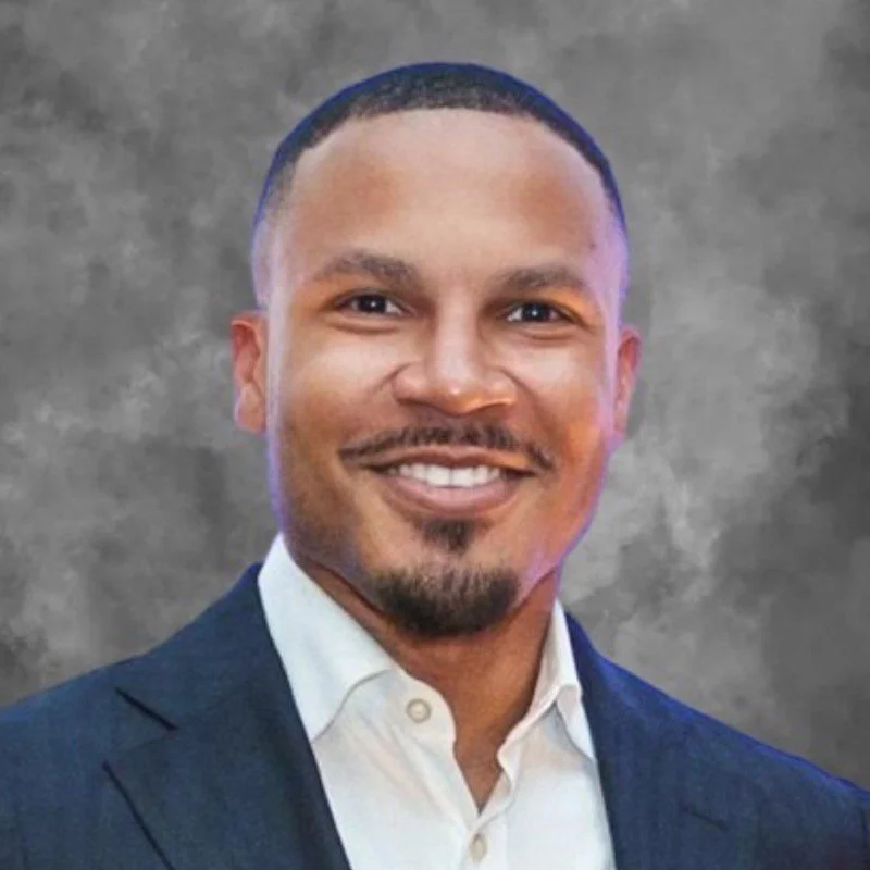Black and white photo of a person wearing glasses, a suit, and a white shirt, smiling in front of a backdrop with text.