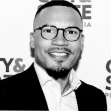 Black and white photo of a person wearing glasses, a suit, and a white shirt, smiling in front of a backdrop with text.