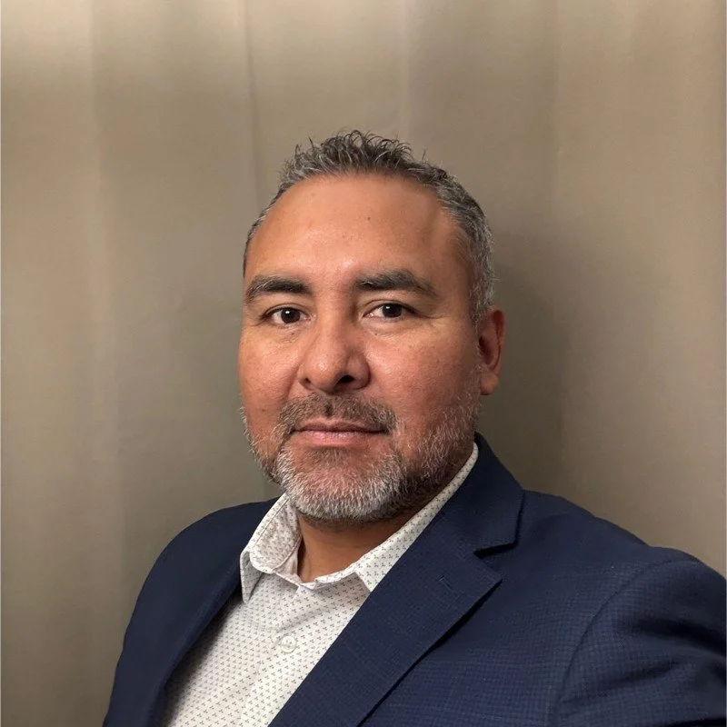 Portrait of a man with a beard wearing a suit jacket, standing against a plain background.