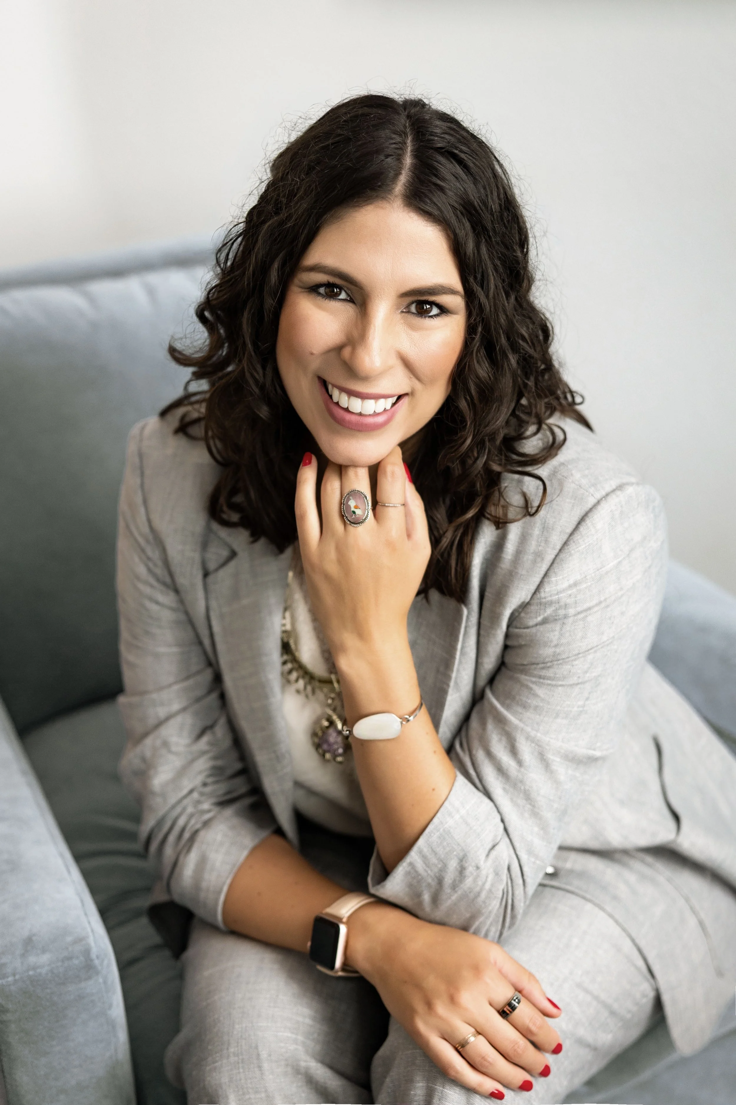 A woman with dark, curly hair sitting on a gray couch, smiling at the camera. She is wearing a light gray blazer and matching pants, accessories including rings, bracelets, and a smartwatch, and has red nail polish.