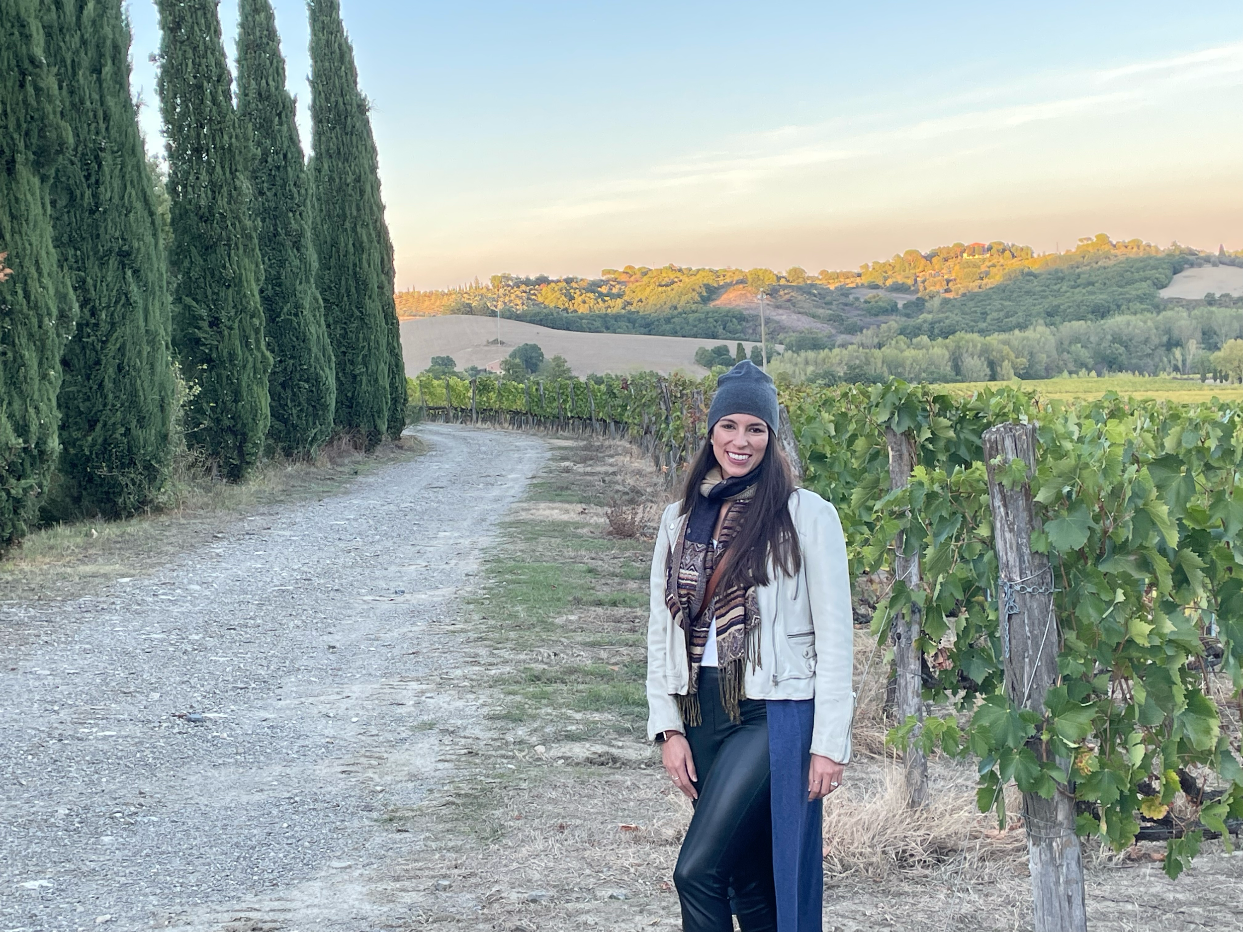 A woman with long brown hair, wearing a gray beanie, white jacket, patterned shirt, and dark pants, standing on a dirt path in a vineyard with green hills and trees in the background, during daylight.