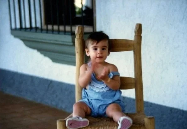 A young child sitting on a wooden chair outdoors, wearing a blue dress and pink shoes.