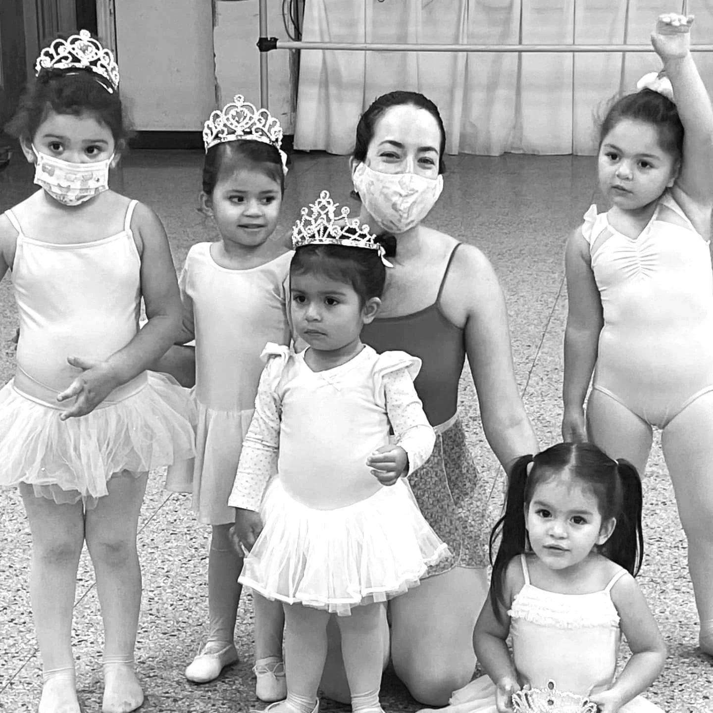 A group of young children dressed in ballet costumes, some wearing tiaras and face masks, with an adult woman in the center also wearing a mask, in a classroom or dance studio setting.
