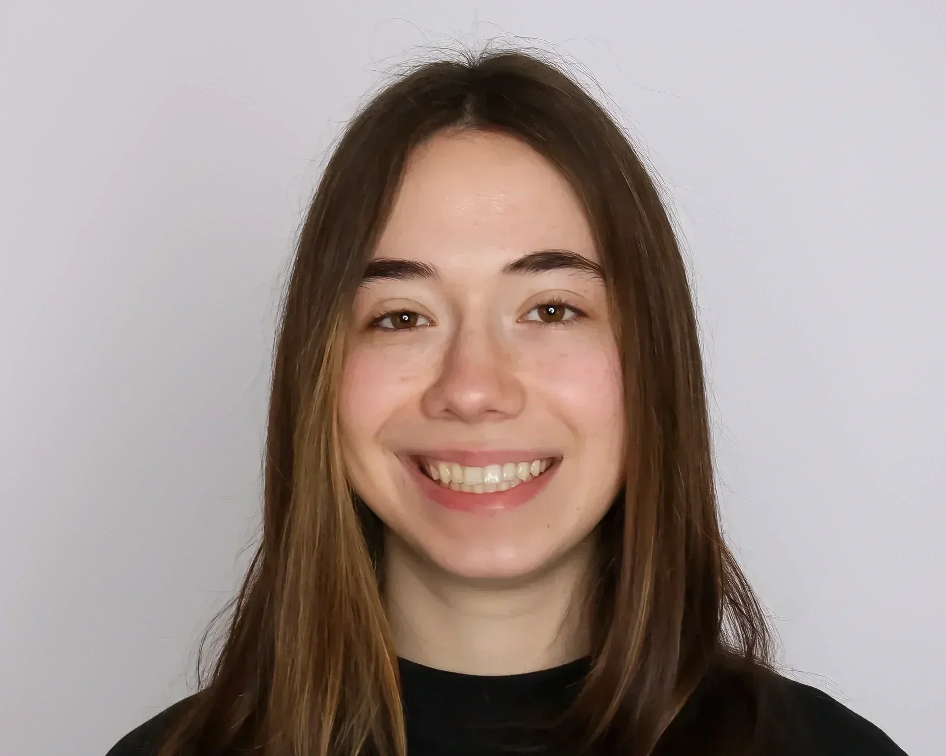 A young woman with long brown hair smiling at the camera against a plain light-colored background.