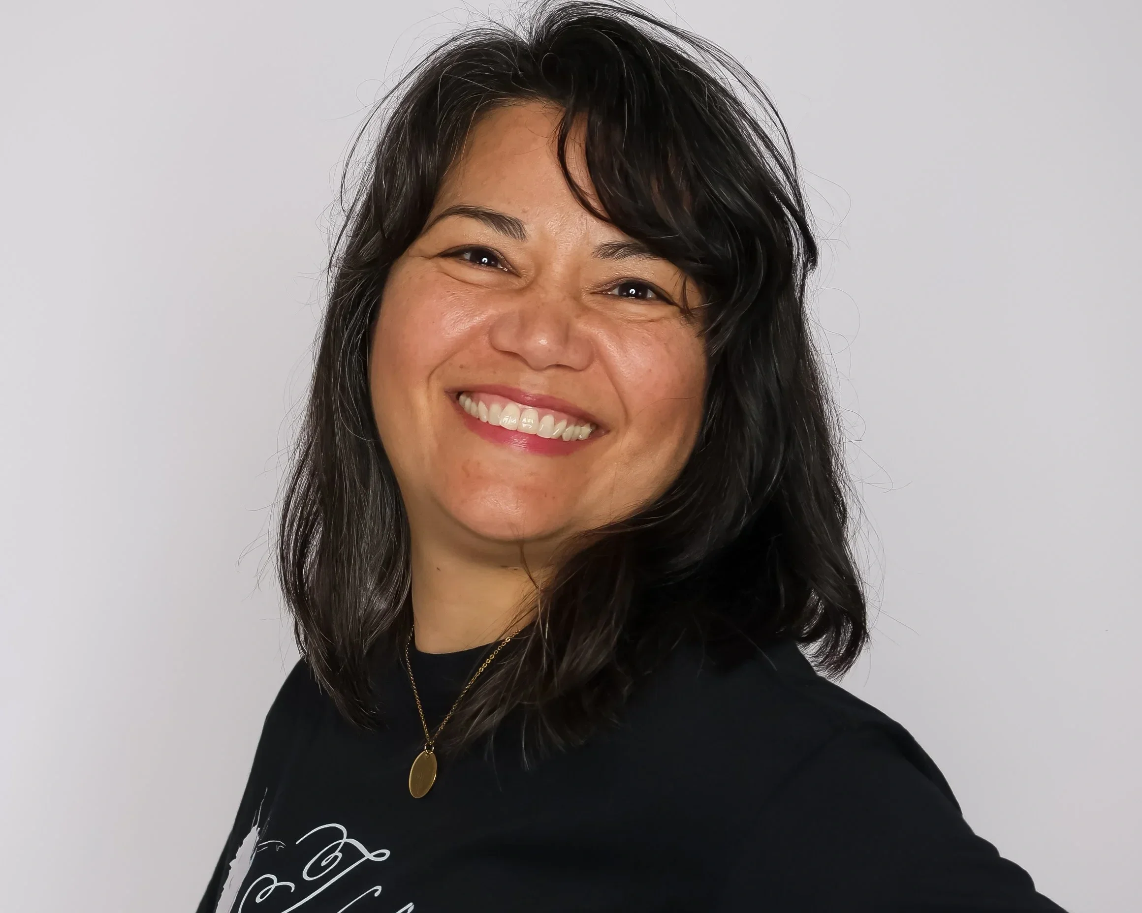 A woman with shoulder-length black hair smiling, wearing a black T-shirt and a gold necklace with a small oval pendant.