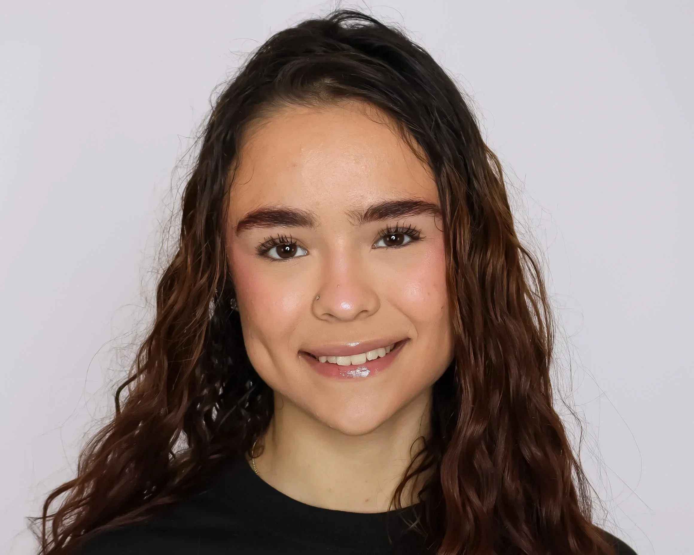 A young woman with long, curly brown hair, smiling, wearing a black top, and a small nose piercing, standing against a plain white background.