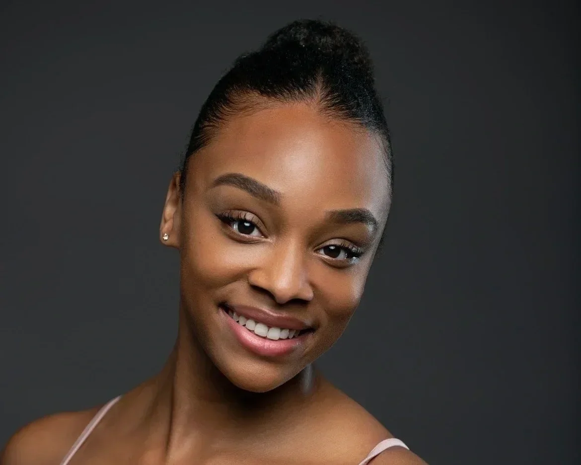 Close-up portrait of a young African American woman smiling, with her hair styled in a bun, wearing small earrings and a light-colored strap top, against a dark background.