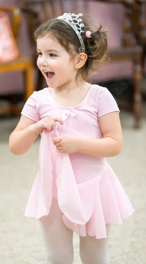 A young girl wearing a pink princess dress with a tiara and a pink hair tie, smiling and holding the fabric of her dress.