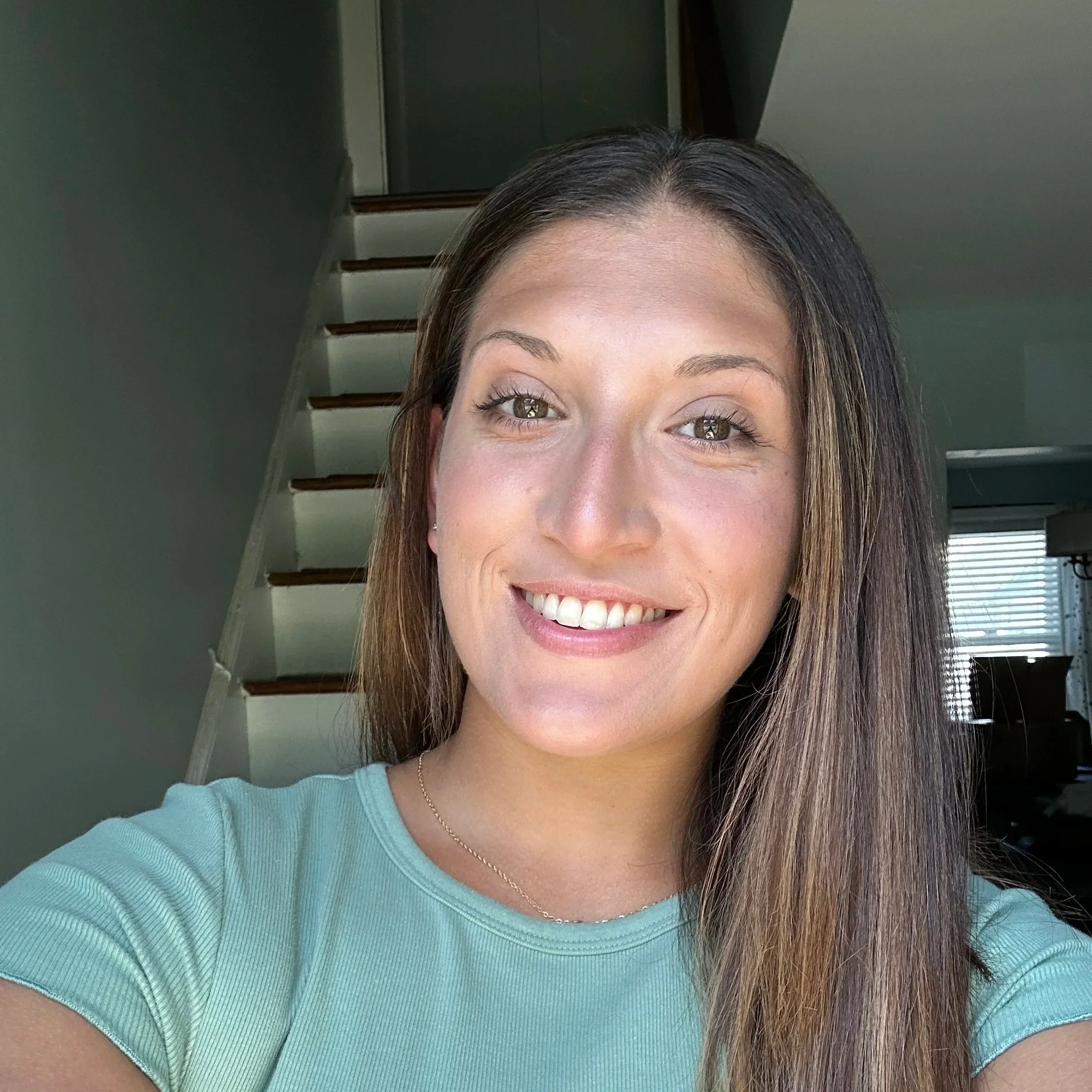Smiling woman with long brown hair taking a selfie indoors with stairs and window in the background.