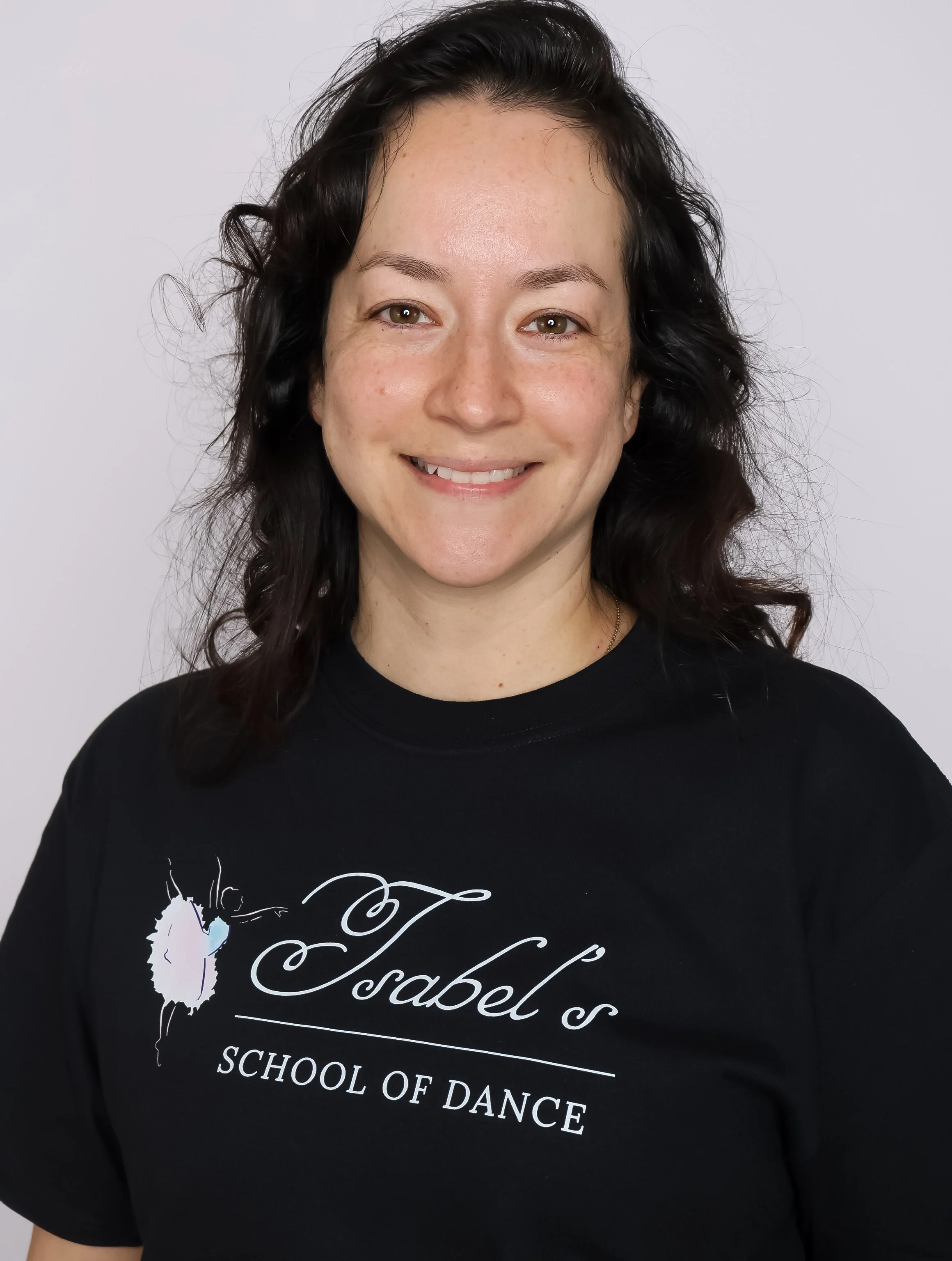 Group of nine women in matching black t-shirts with 'Isabel's School of Dance' logo, posing in front of a plain gray background. Some are kneeling, others standing, smiling at the camera.