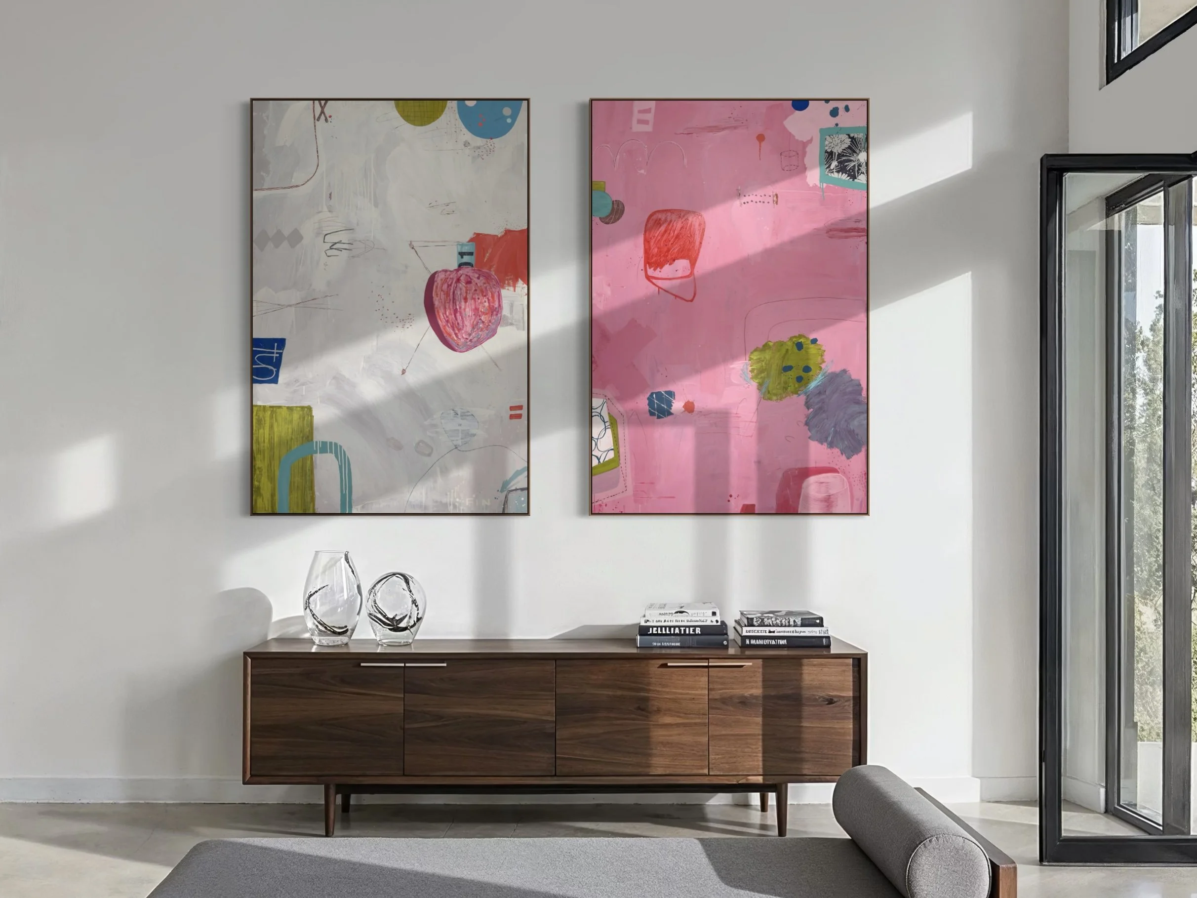 Interior view of a modern living room with two large abstract paintings hanging on a white wall, a wooden sideboard with decorative glass vases and books, and a gray sectional sofa in the foreground near large windows letting in natural light.