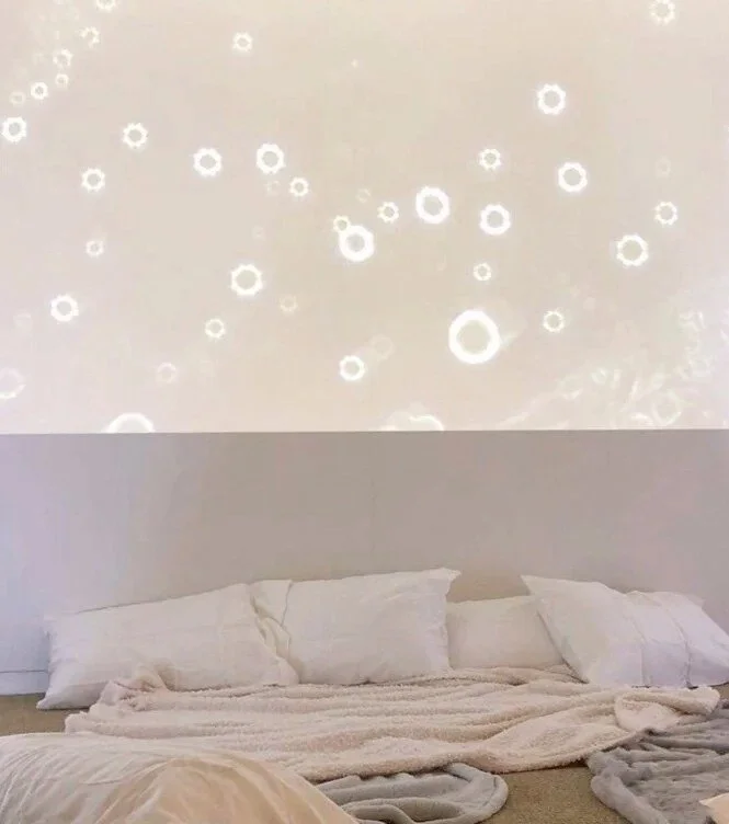 Reflections of Light on Breastmilk Particles, 2018, by Eva Zasloff and Laura Zittrain. Glowing rings of light projected on a wall above a bed of white pillows and blankets at the MIT Media Lab.