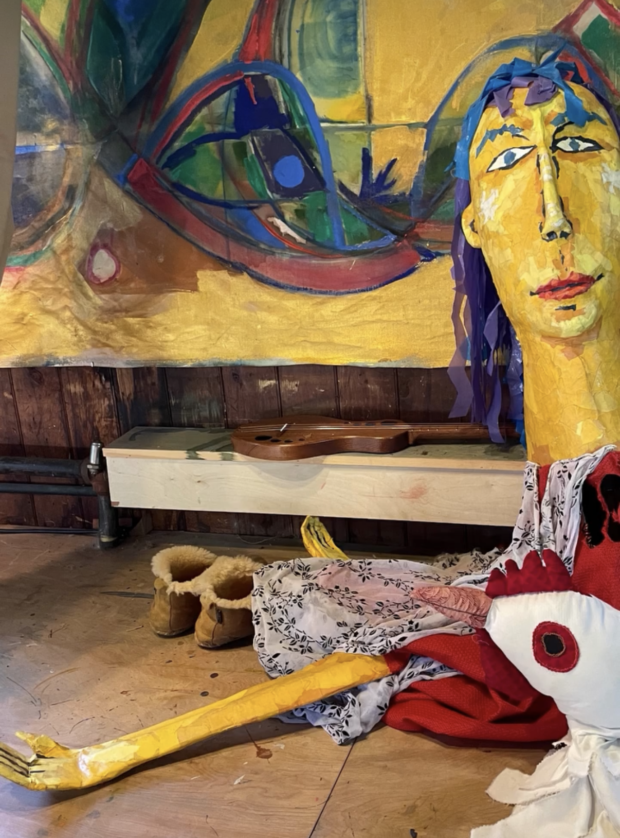 A large papier-mâché puppet with a yellow face and long yellow arm, on loan from the Free Puppet Library, resting on the floor of the Barn at Schwamb Mill beneath a painting by Eva Zasloff, for performances of Ruth Krauss and Maurice Sendak's A Very 