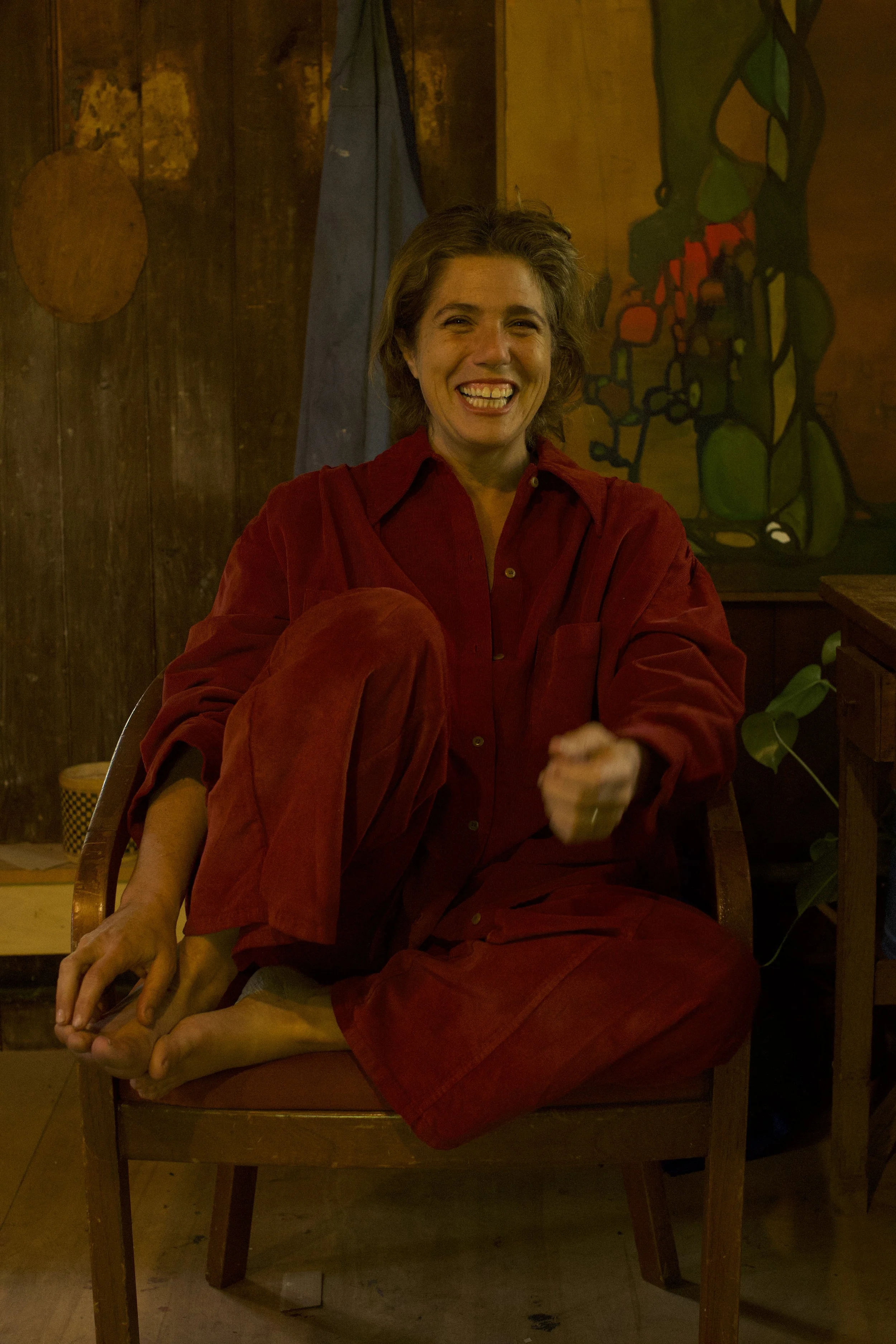 Eva Zasloff laughing, seated in a red corduroy suit in her barn studio, with one of her paintings behind her.