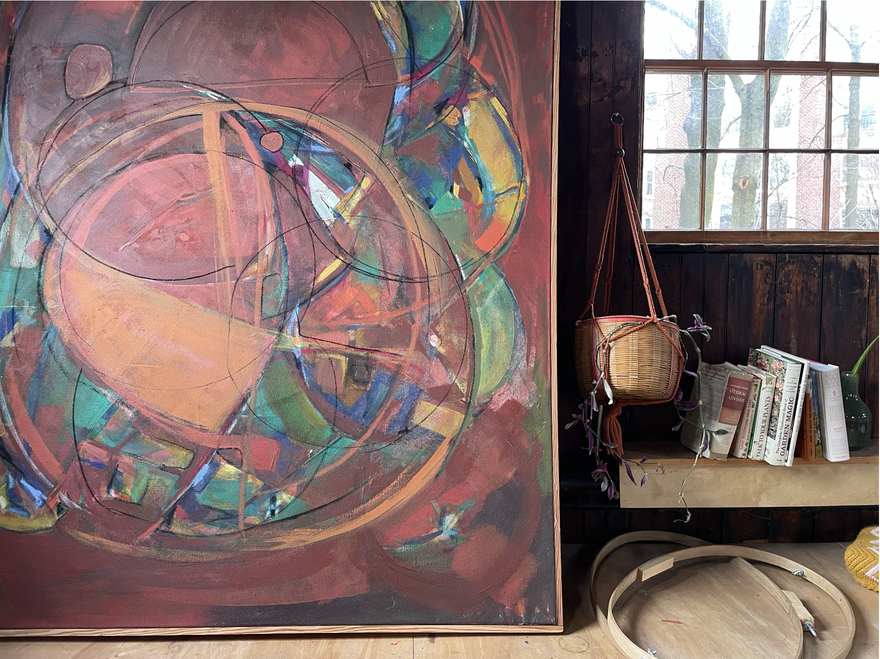 An abstract colorful painting with circular and geometric shapes on a wall, next to a window and shelves with books and a hanging wicker basket.