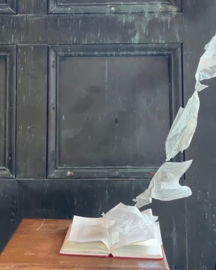 Air Breathing by Eva Zasloff, 2024: pages from a 1964 medical textbook on the newborn's adaptation to life outside the womb rise from an open book on a wooden table, suspended in the air against a dark door. Shown in Rebirth Reimagine, The Substation