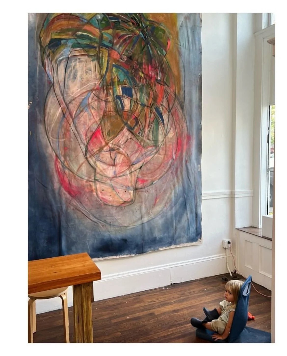 An abstract painting with bright colors and energetic lines hung on a white wall in a room with wooden flooring. A small child sitting in a blue chair in front of the painting, looking at it.