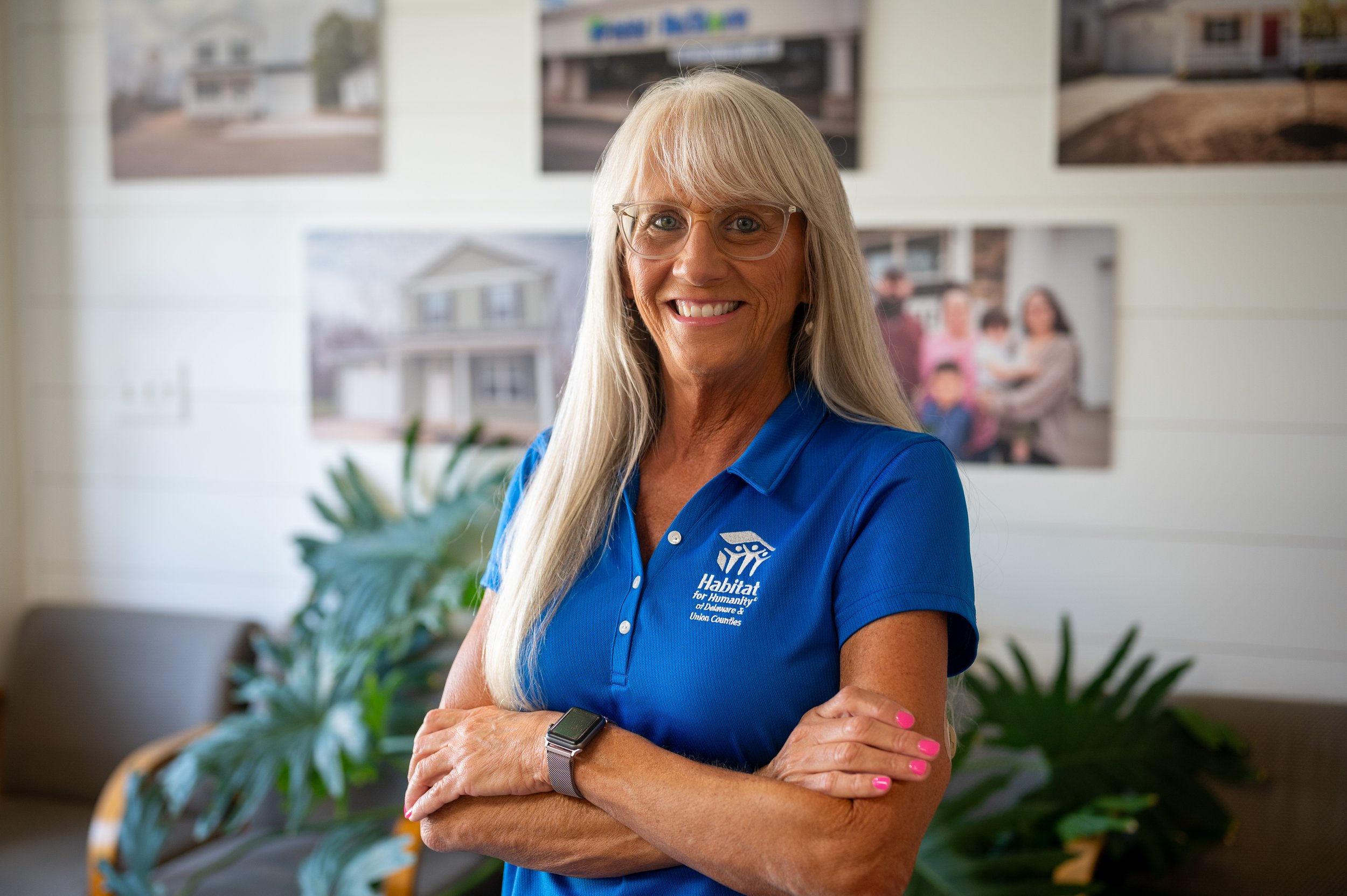 A woman wearing glasses and a blue jacket with a Habitat for Humanity logo, smiling at a construction site with wooden framing in the background.