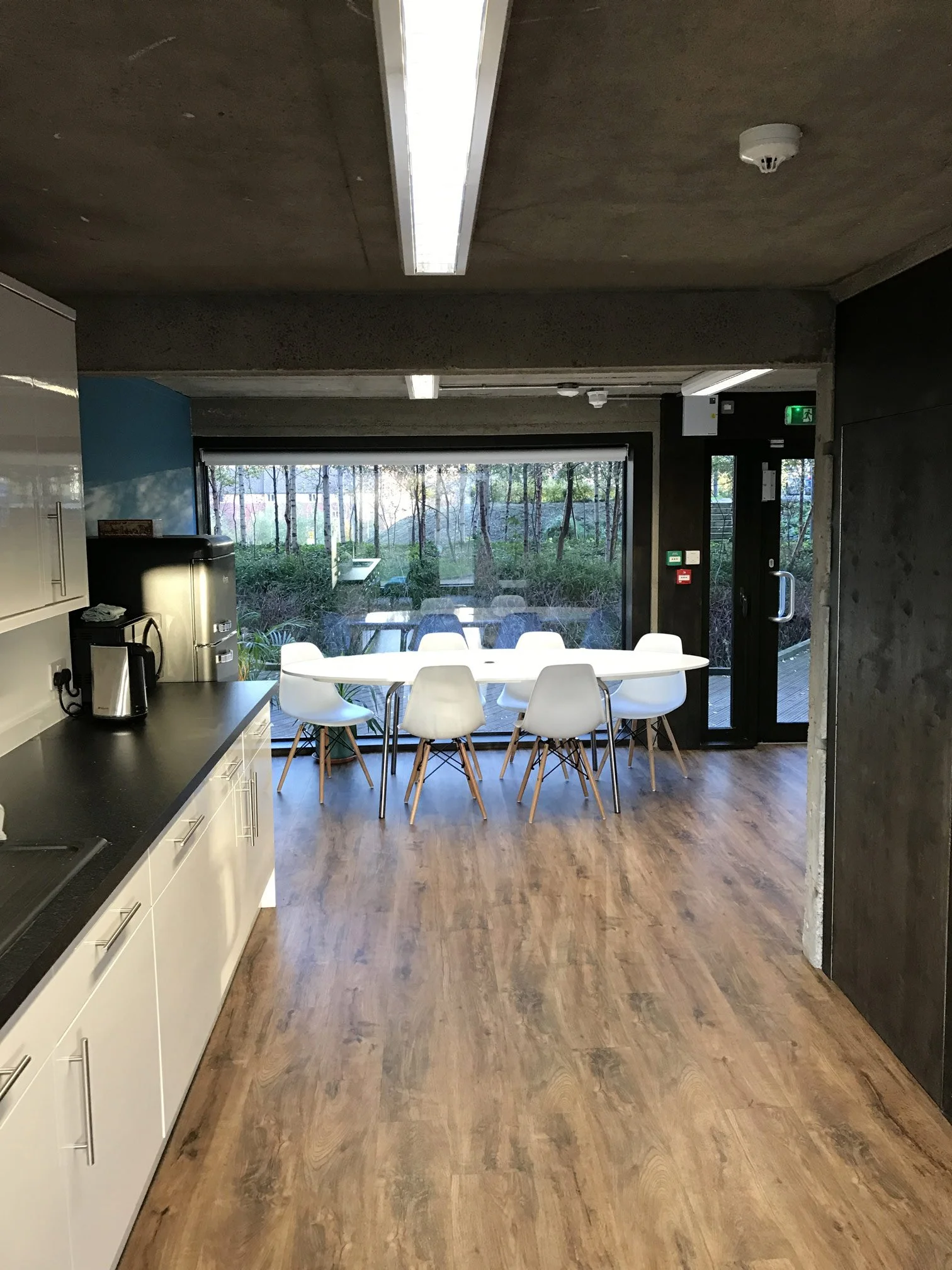 Interior view of a meeting room with a white round table surrounded by white chairs, large window revealing trees outside, and kitchen counter with coffee maker, in a modern office.