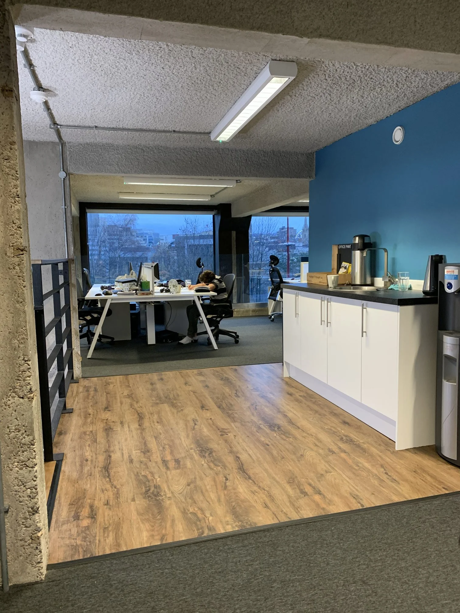 An office breakroom with a blue accent wall, wooden floor, white cabinets, and a countertop with a coffee urn, kettle, and cups. There is a kitchenette area with a large window showing an urban cityscape outside. The ceiling has textured white panels