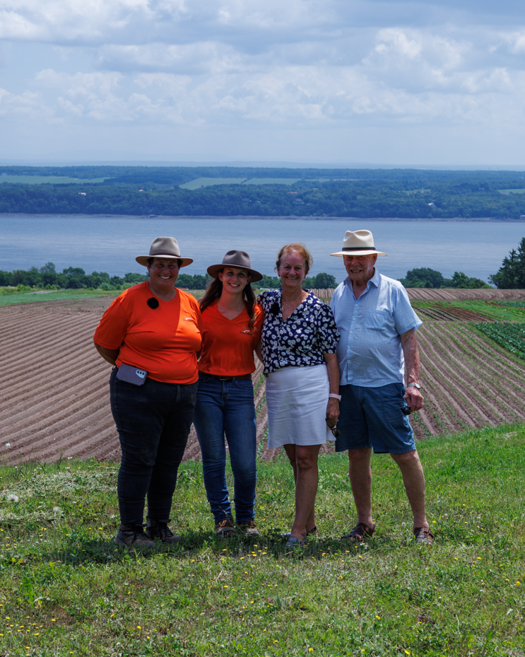 Quatre personnes se tenant côte à côte dans un champ, avec un lac et une plaine verte en arrière-plan, toutes portant des chapeaux et souriant.