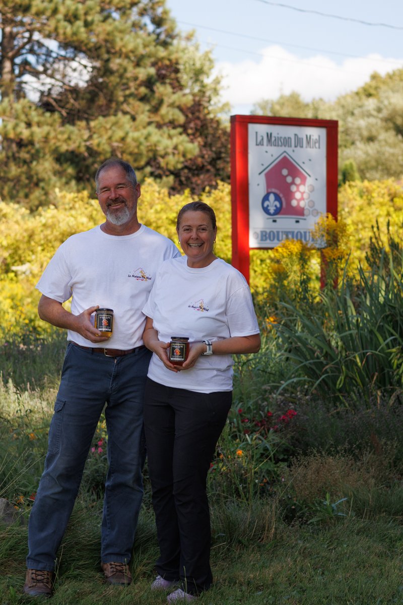 Deux personnes souriantes, un homme et une femme, tenant des pots de miel, se tenant devant un panneau indiquant 'La Maison Du Miel' et un environnement naturel avec des arbres et des fleurs.