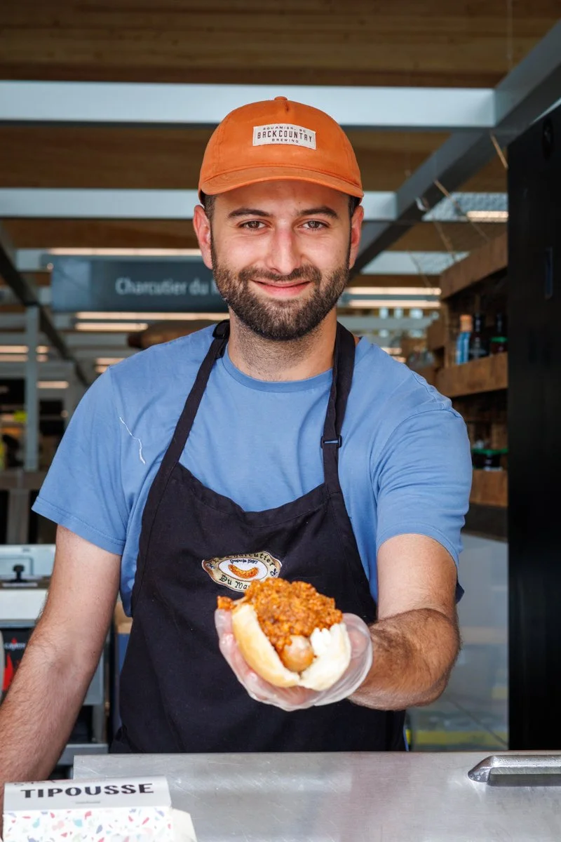 Homme souriant portant une casquette orange et un tablier noir, tenant un hot-dog avec de la viande hachée ou du chili, à l'intérieur d'une maison ou d'un restaurant.