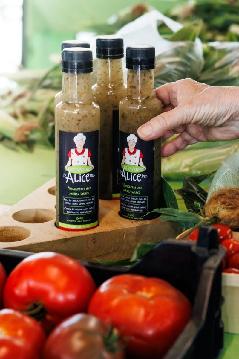 Bouteilles de sauce assaisonnement à l'ail avec une illustration d'une femme chef sur l'étiquette, posées sur un support en bois dans un marché de produits frais, avec des tomates en premier plan.