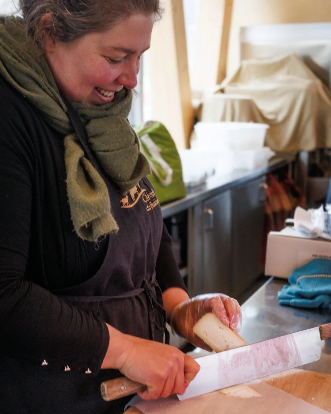 Une femme souriante coupe du fromage avec un couteau dans une cuisine.