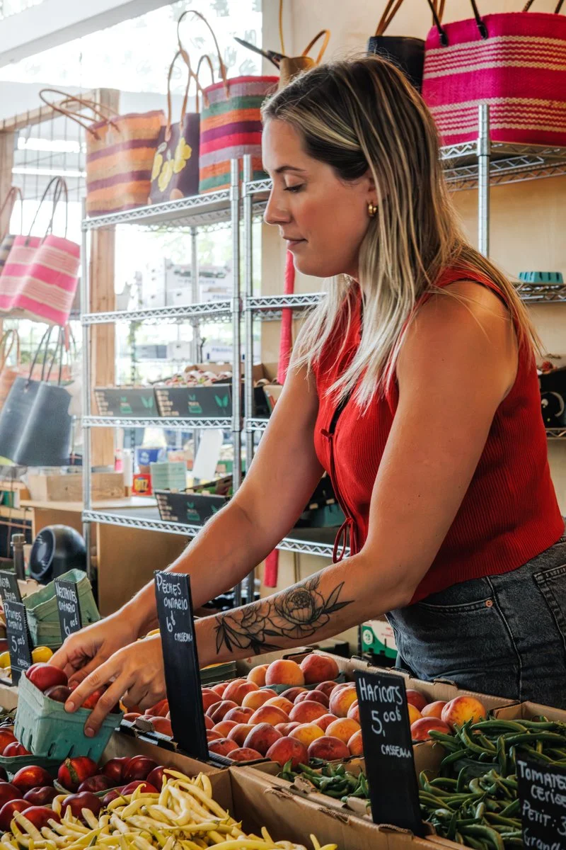 Une femme achetant des fruits au marché, entourée de paniers en bandoulière suspendus. Elle porte un haut rouge et a des tatouages sur le bras.
