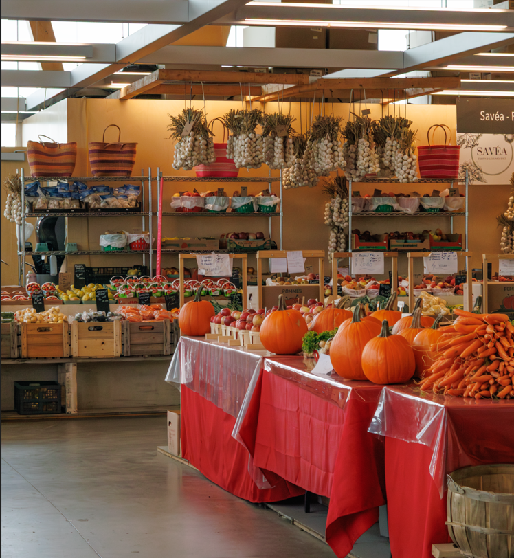 Stand de légumes et de citrouilles dans un marché couvert, avec des carottes, des pommes et des filets d'ail suspendus.