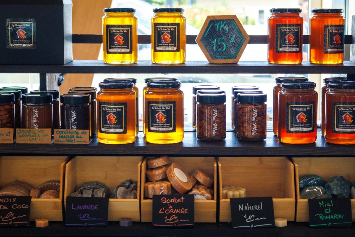 Étagère avec des pots de miel, des savons artisanaux, et des étiquettes avec des noms de parfums comme lavande, orange, nature, miel et romarin.