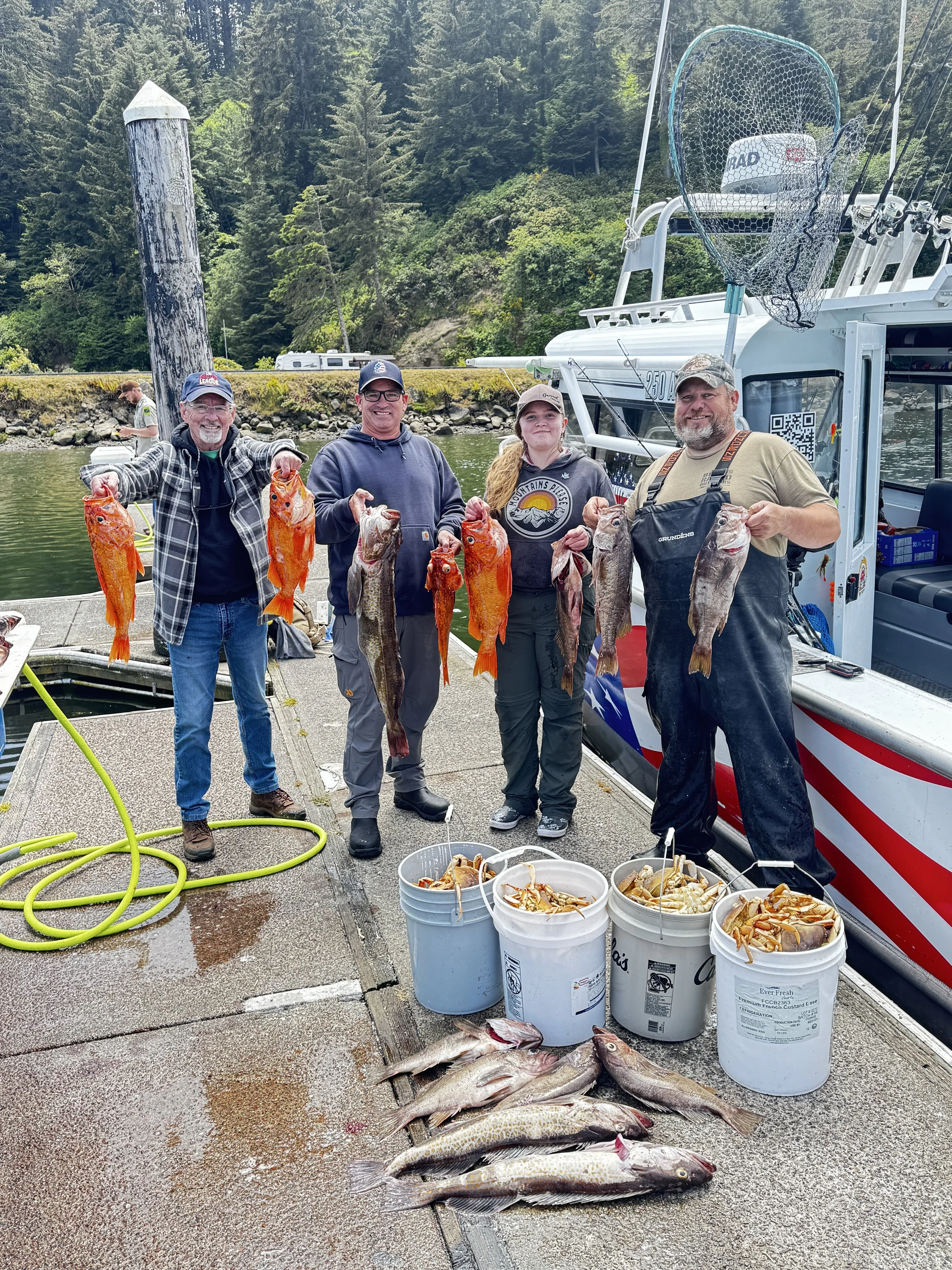 Four people standing on a dock holding fish and crabs after a fishing trip, with buckets of caught seafood in front of them, a boat behind, and a background of trees and water.