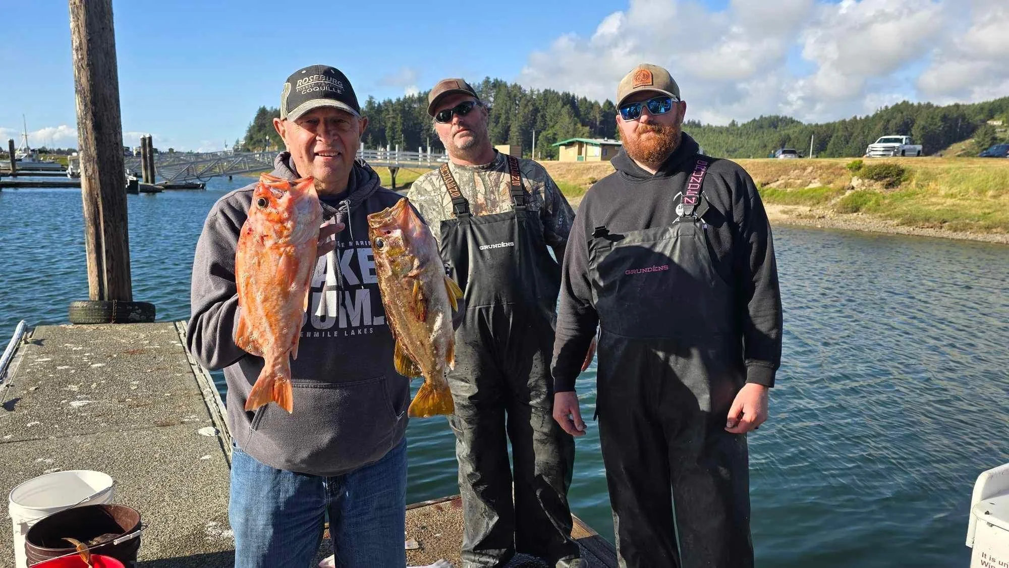 Three men standing on a dock by the water, two holding fish, wearing outdoor clothing and hats, with a background of trees and a partly cloudy sky.