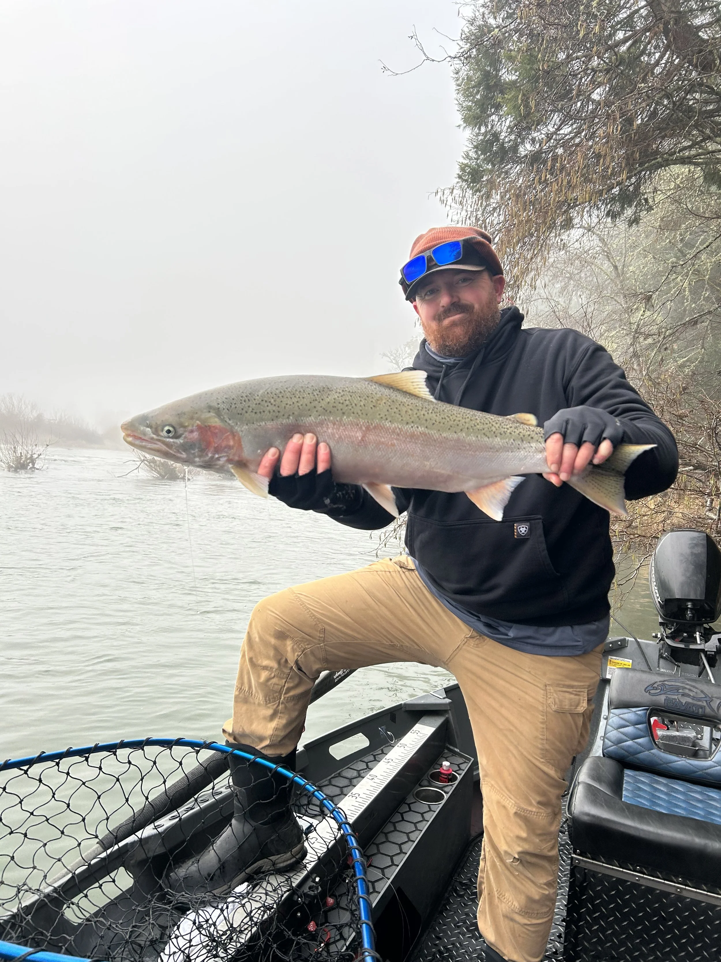 A man with a beard, wearing a brown beanie, sunglasses, a black jacket, and tan pants, is holding a large fish with both hands while on a boat in a foggy lake. The fish is a rainbow trout, and there is fishing gear and a measuring ruler on the boat.