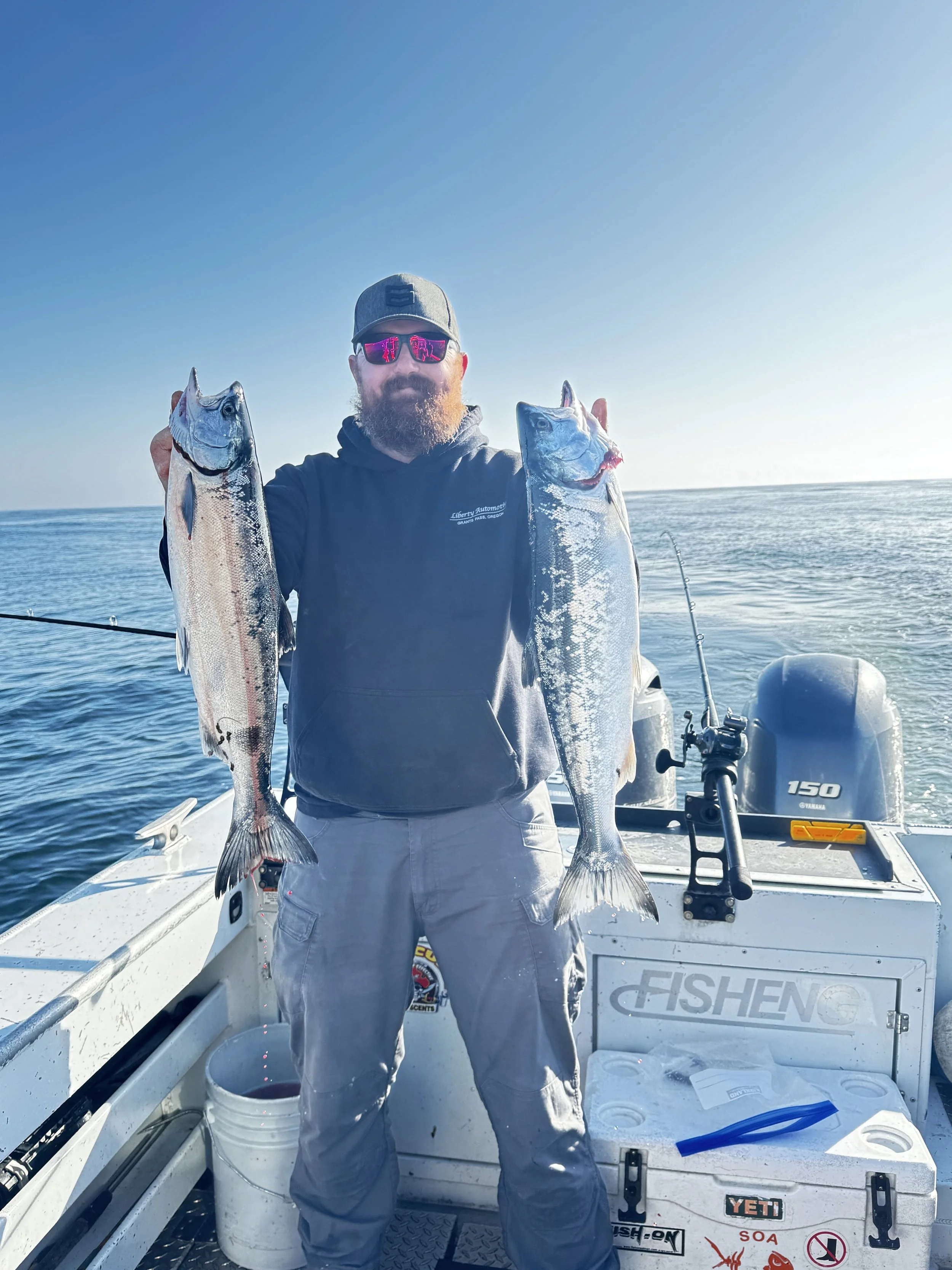 A bearded man wearing sunglasses and a gray cap holding two large fish on a boat in the ocean during daylight.