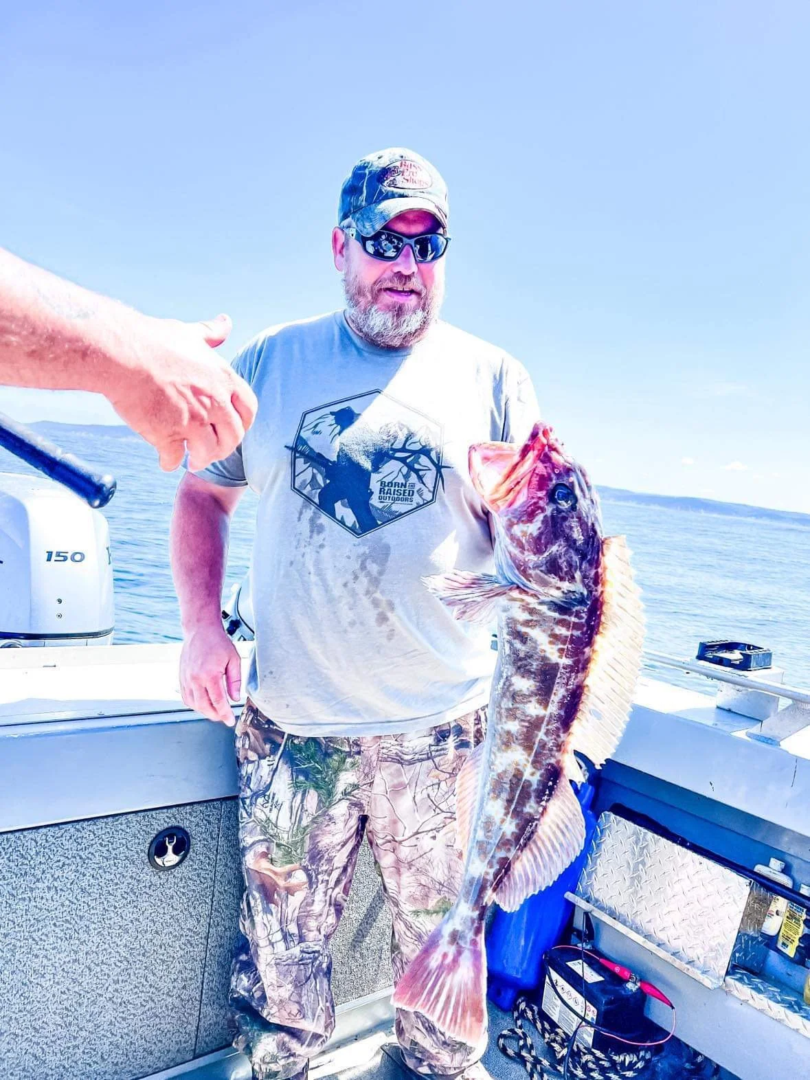 A person wearing sunglasses and camo clothing holding a large fish on a boat with a blue ocean in the background.