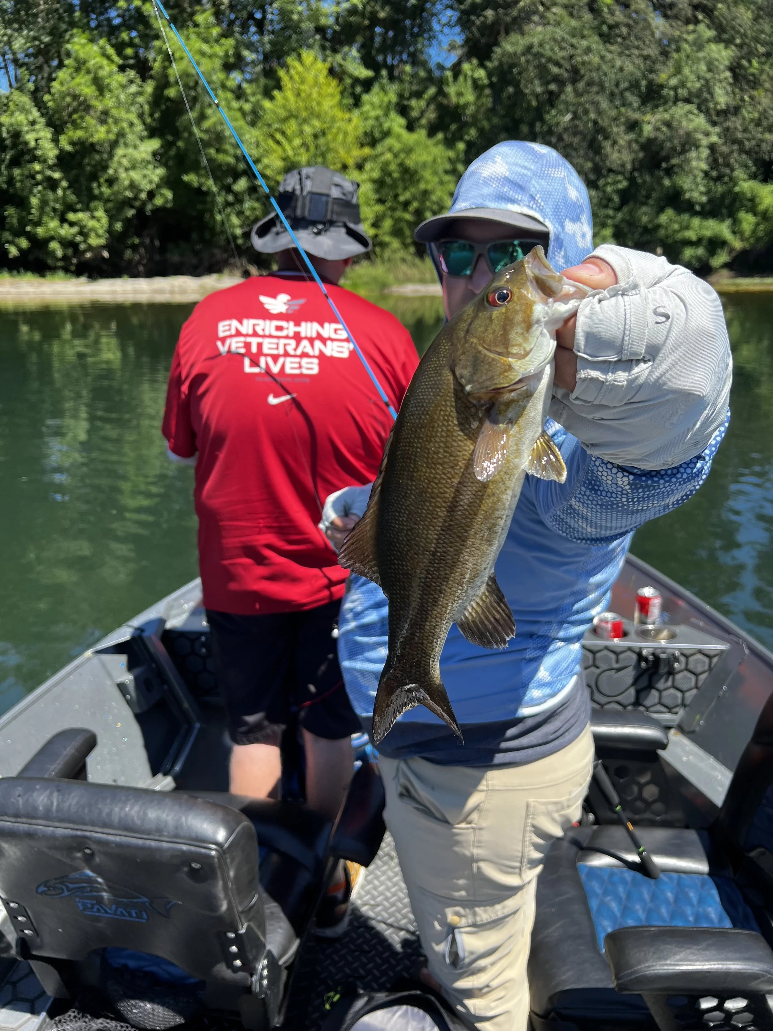 Two people fishing on a boat, one holding a fish. One person is wearing a red shirt with text, the other is wearing a blue jacket and sunglasses. Trees and water in the background.