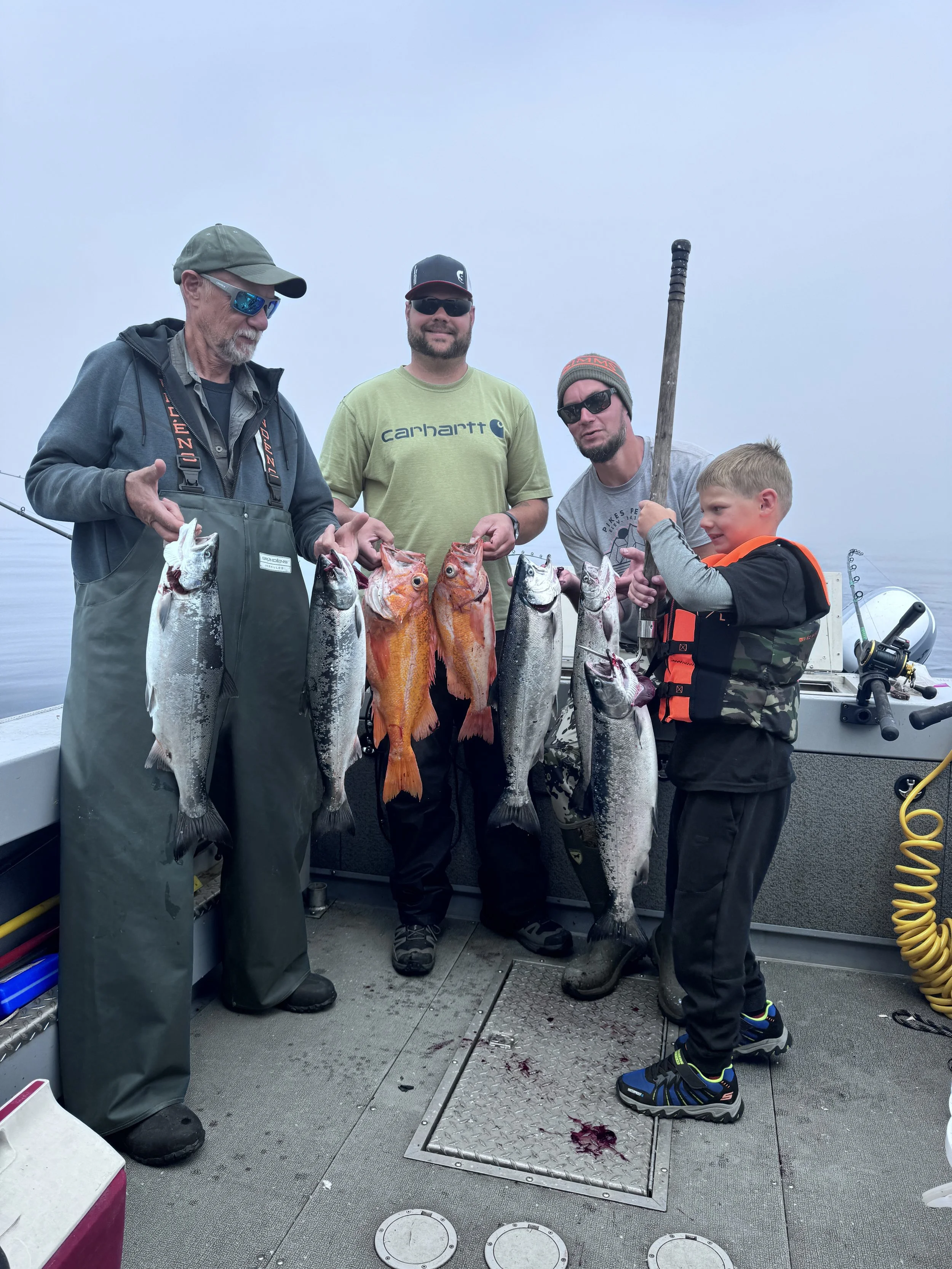 Group of people on a boat holding freshly caught fish, including salmon and rockfish, with one child holding a fishing gaff. The boat floor is visible with fishing equipment around.