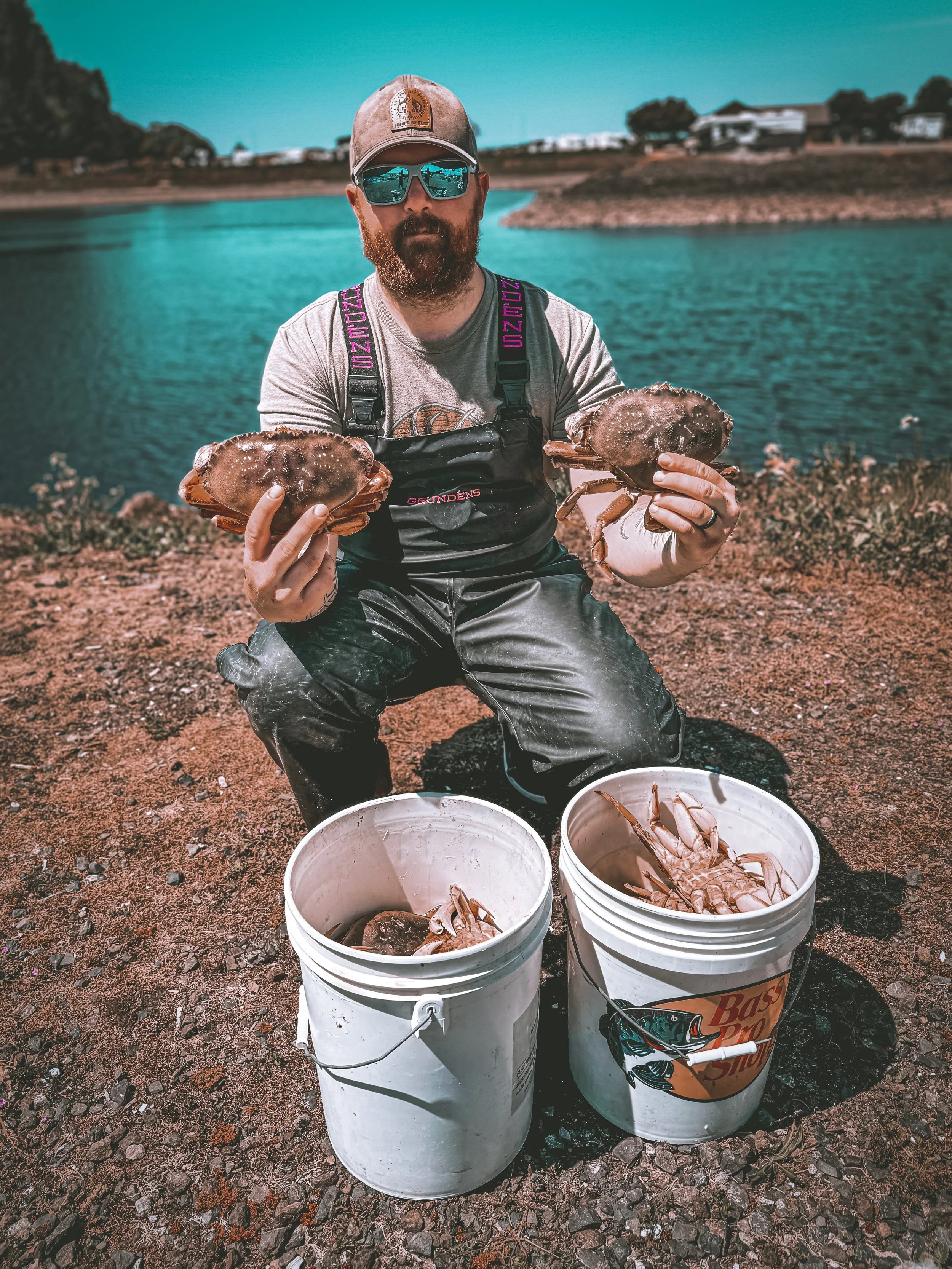 A person wearing a cap and sunglasses squatting near a body of water, holding two crabs. Two buckets filled with crabs are on the ground in front of them.